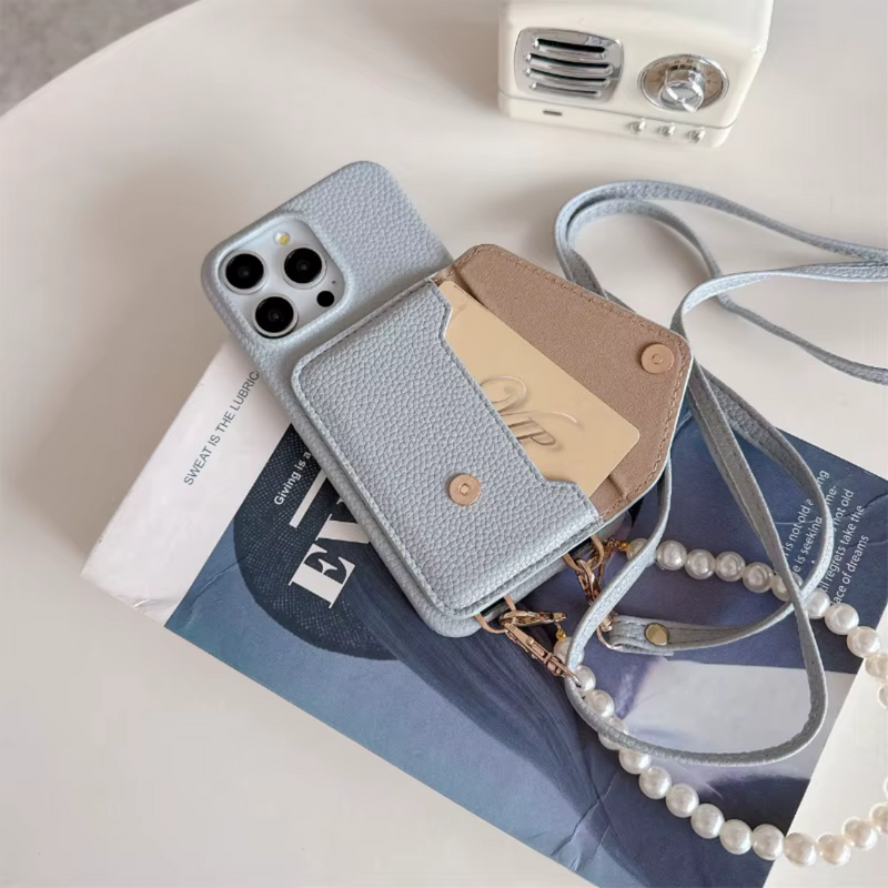 iPhone Umhängehülle Pearl in Hellblau aus veganem Leder mit geöffnetem Kartenfach, Perlenkette und verstellbarem Umhängeband – stilvoll arrangiert auf einem Magazin mit dekorativem Mini-Lautsprecher.